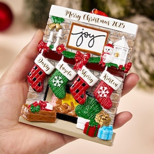 May include: A Christmas-themed ornament featuring a fireplace design with stockings labeled with names. The ornament includes the text "Merry Christmas 2025" and the word "joy". Red, green, and white colors are prominent.