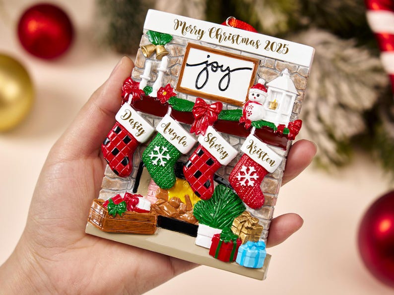 May include: A Christmas-themed ornament featuring a fireplace design with stockings labeled with names. The ornament includes the text "Merry Christmas 2025" and the word "joy". Red, green, and white colors are prominent.