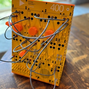 May include: A yellow and orange pocket operator modular synthesizer with gray patch cables connected to the various inputs and outputs. The synthesizer has a label that reads "pocket operator™ modular" and "400."