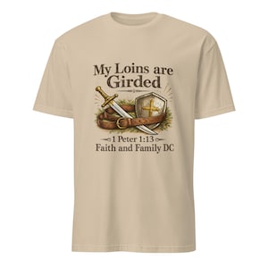 Puede incluir: Camiseta beige con un diseño gráfico de espada, escudo y cinturón. El texto dice "My Loins are Girded" con "1 Peter 1:13 Faith and Family DC" debajo. El diseño utiliza colores marrón, dorado y verde.