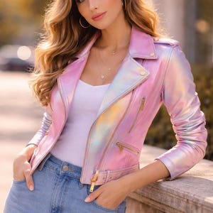 Puede incluir: Chaqueta de motorista rosa iridiscente con cremalleras y herrajes dorados. La chaqueta tiene un diseño asimétrico clásico con cuello de solapa y cintura con cinturón. La modelo lleva un top blanco y vaqueros azules.