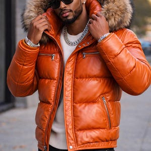 May include: A tan leather puffer jacket with a fur-trimmed hood. The jacket features a zip closure, multiple pockets, and a quilted pattern. The person in the image is wearing sunglasses and a silver chain necklace.
