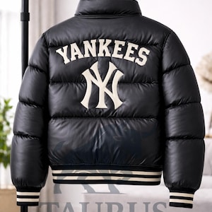 Op de afbeelding: Een zwarte gewatteerde jas met het woord YANKEES in witte letters boven een groot wit NY-logo op de achterkant. De jas heeft een hoge kraag, lange mouwen met geribbelde boorden en een geribbelde zoom met witte strepen. Het materiaal glanst licht.