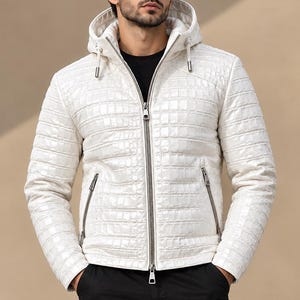 Pode incluir: Um casaco branco com capuz e textura de pele de crocodilo, com fecho de correr prateado a todo o comprimento e dois bolsos laterais com fecho. O casaco é usado sobre uma t-shirt preta, combinado com calças cargo pretas e óculos de sol escuros.