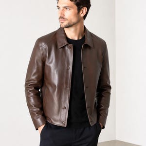 Könnte beinhalten: Braune Lederjacke mit Knopfleiste und Kragen. Die Jacke hat eine mittelbraune Farbe und scheint aus hochwertigem Leder gefertigt zu sein. Sie wird über einem schwarzen Hemd getragen.