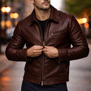 Könnte beinhalten: Braune Lederjacke mit Reißverschluss und Kragen. Die Jacke hat eine kleine Brusttasche und wird über einem schwarzen Shirt getragen. Das Model trägt dunkelblaue Jeans.
