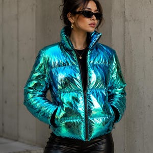 Op de afbeelding: Een metallic, iriserend gewatteerd jack met een hoge kraag, lange mouwen en een ritssluiting aan de voorzijde. Het jack heeft een levendige, reflecterende blauwe en groene afwerking met een doorgestikte textuur, gedragen over een zwarte top.