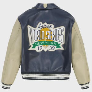 May include: A navy blue and beige leather jacket featuring the text "Avirex WORLD SERIES NEW YORK 1939" on the back. The jacket has a ribbed hem and cuffs with white stripes. The sleeves are beige, and the body is navy blue.