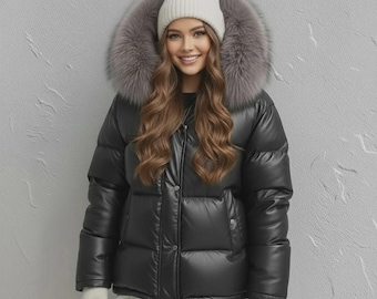 May include: A black puffer jacket with a fur-trimmed hood and a white beanie. The jacket has a glossy finish and a button-up closure. The fur trim is gray. The person is wearing white gloves.