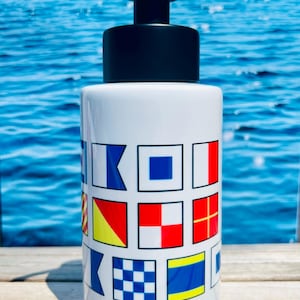 May include: White ceramic soap dispenser with a black pump. The dispenser is decorated with colorful nautical signal flags. The background is a blurred image of the ocean.