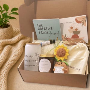 Könnte beinhalten: Eine Geschenkbox mit Artikeln zur Selbstpflege. Enthält ein Journal, ein Malbuch, eine Kerze, einen Becher, eine gehäkelte Sonnenblume und eine kleine Häkelfigur mit einem Schild "Thank you for being Awesome."