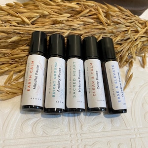 May include: Five essential oil roller bottles with black caps and white labels. The labels read "Earth Calm," "Bright Ground," "Sacred Heart," "Clear & Calm," and "Ease & Elevate." The background includes dried wheat stalks.