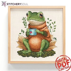 May include: Cross-stitch pattern of a green frog wearing an orange sweater and holding a blue mug with a pink heart. The frog is sitting on a leaf, with green foliage around it. The framed artwork is labeled "DIGITAL PDF PATTERN".