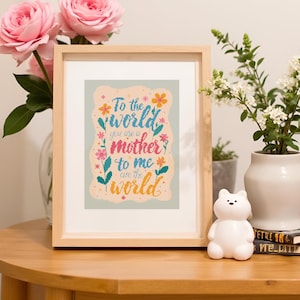 以下が含まれることがあります： 「To the world you are a mother to me are the world」というカラフルな文字が書かれた、額入りのクロスステッチアート。 フレームは明るい木製で、アートワークは花で飾られています。 作品は木製のテーブルに飾られています。