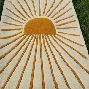 Puede incluir: Una alfombra rectangular con un diseño de rayos de sol. La alfombra presenta un fondo color crema con un gran sol amarillo dorado en el centro. Los rayos amarillo dorado se extienden hacia afuera desde el sol, creando un efecto visual llamativo.
