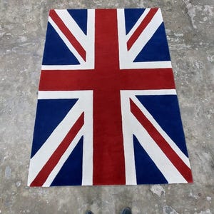 Puede incluir: Una alfombra rectangular con la bandera del Reino Unido, la Union Jack. El diseño incluye una cruz roja y rayas diagonales sobre un fondo azul, con detalles blancos. La alfombra está colocada sobre una superficie de hormigón.