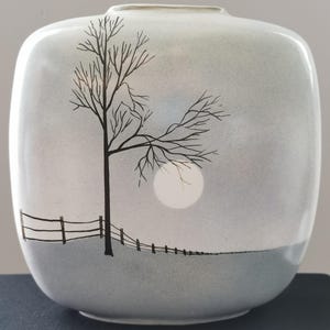 Könnte beinhalten: Eine Keramikvase mit Landschaftsdesign. Die Vase zeigt einen schwarzen Baum, einen Zaun und einen weißen Mond vor grauem Hintergrund. Der Boden der Vase ist dunkelgrau.