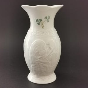 May include: A white porcelain vase featuring a flared rim and a tapered neck. The body is decorated with embossed relief carvings of figures and intricate knotwork patterns, accented by a small, painted green shamrock motif near the top.
