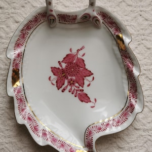 May include: White porcelain leaf-shaped serving tray with a handle. The tray features a pink floral design in the center and a pink and gold border. Gold accents are placed on the rim and handle.