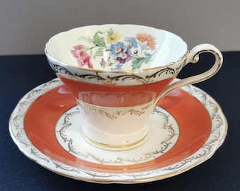 May include: A vintage teacup and saucer set. The cup and saucer are white with a coral band and gold trim. The cup features a floral design with pink, blue, and yellow flowers. The set is likely made of porcelain.
