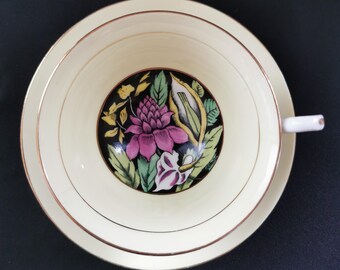 May include: Cream-colored teacup and saucer set with a floral design. The cup's interior features a black circle with a pink flower and green leaves. Gold trim accents the edges of the cup and saucer. The handle is white.