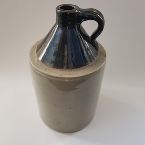May include: A stoneware jug featuring a cylindrical body with a smooth, light beige finish and a contrasting dark, glossy blue-black glazed neck. The vessel includes a single curved handle attached to the upper neck and a narrow circular opening.