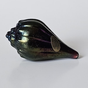 May include: A dark, iridescent glass seashell sculpture with a tapered, pointed end and a rounded, spiraled base. The surface features deep green and purple hues with a metallic sheen. An oval sticker reads: ROBERT HELD HAND MADE IN CANADA ART GLASS.