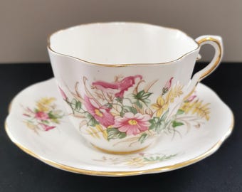May include: White teacup and saucer set with a floral design featuring pink and yellow flowers and green leaves. The cup and saucer have gold trim along the edges. The teacup has a curved handle.