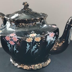 May include: A black ceramic teapot with a lid, handle, and spout. The teapot is decorated with pink and yellow floral designs and gold trim. The teapot is a vintage style and is likely used for serving tea.