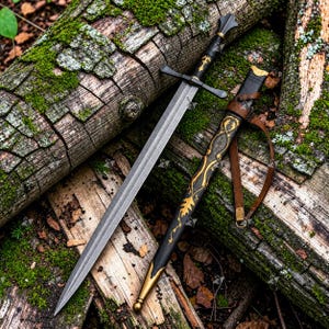 May include: A double-edged steel sword with a black hilt and a crossguard. Beside it lies a black scabbard featuring gold leaf-patterned filigree, a gold-colored metal tip, and a brown leather strap for carrying the weapon securely.