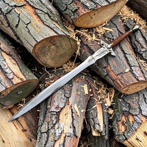 May include: A silver-toned metal sword with a pointed blade featuring a central fuller and an intricate crossguard. The handle is made of dark brown wood with a textured pattern and a metallic pommel. The blade is engraved with the text My blade is your brother.