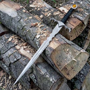 May include: A steel sword with a wavy, flame-bladed edge, a silver crossguard with curved quillons, a black wrapped handle, and a circular pommel featuring an embedded amber-coloured gemstone at the end of the hilt.