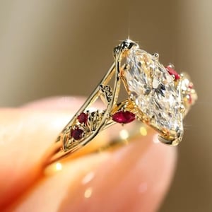 以下が含まれることがあります： 大きなマーキス型の透明な宝石を中央に配したゴールドカラーのリング。リングは、小さな赤い宝石と、バンドに施された複雑なフィリグリーのディテールで装飾されています。リングはヴィンテージなデザインです。