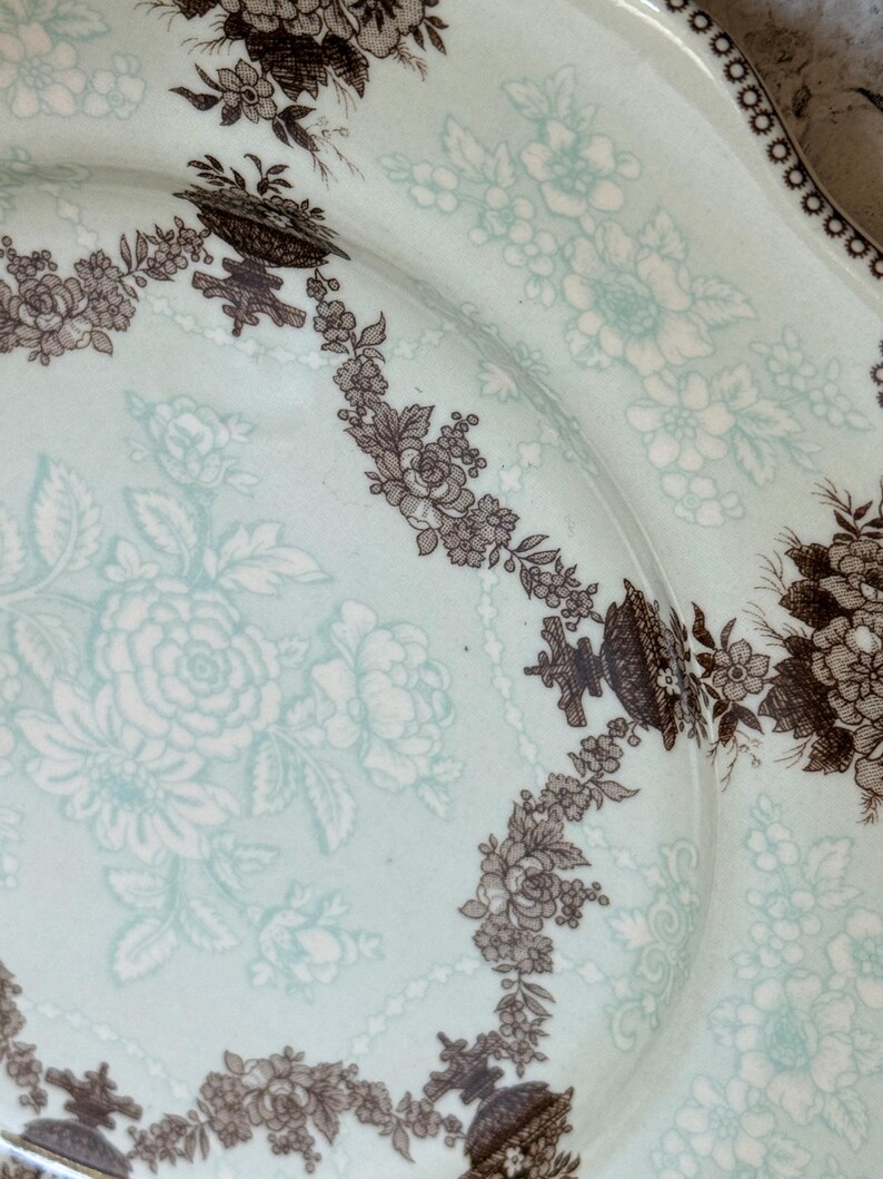 May include: A vintage ceramic plate with a light blue background and a floral pattern in brown and white. The plate features a detailed border design and a delicate floral motif throughout the center.