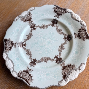 May include: A decorative plate with a scalloped edge. The plate features a light blue background with a floral pattern in white and brown. The rim has a white, textured design.