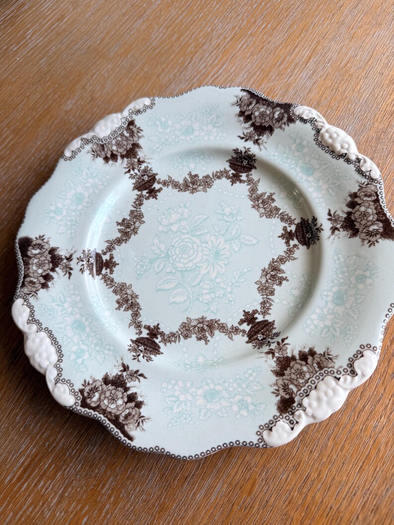 May include: A decorative plate with a scalloped edge. The plate features a light blue background with a floral pattern in white and brown. The rim has a white, textured design.
