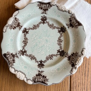 May include: A vintage-style plate with a light blue background and a floral pattern in white and brown. The plate has a scalloped edge with a white and brown trim. The plate is likely ceramic and suitable for serving food.