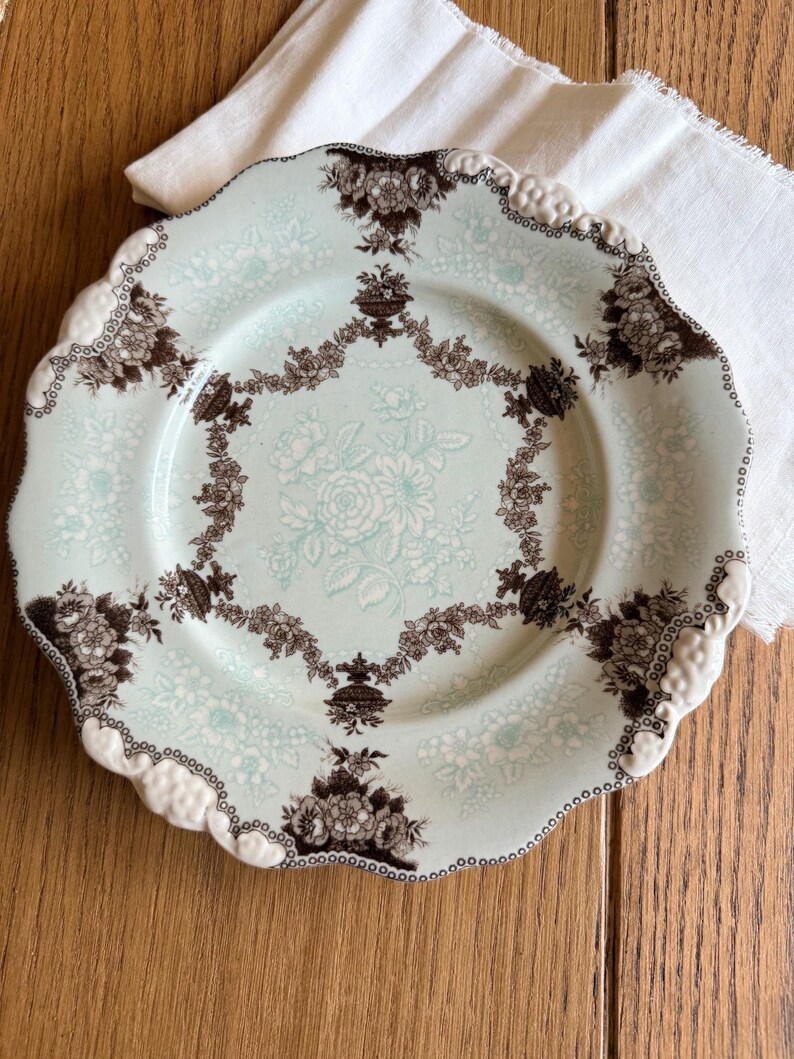 May include: A vintage-style plate with a light blue background and a floral pattern in white and brown. The plate has a scalloped edge with a white and brown trim. The plate is likely ceramic and suitable for serving food.