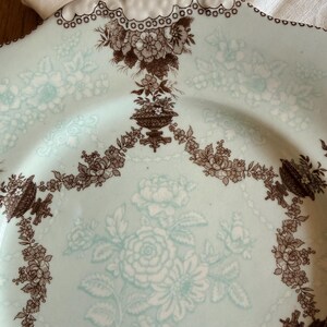 May include: A vintage, light blue plate with a floral design. The plate features a brown floral pattern around the edges and in the center. The rim has a scalloped edge with a white trim. The plate is likely made of ceramic.