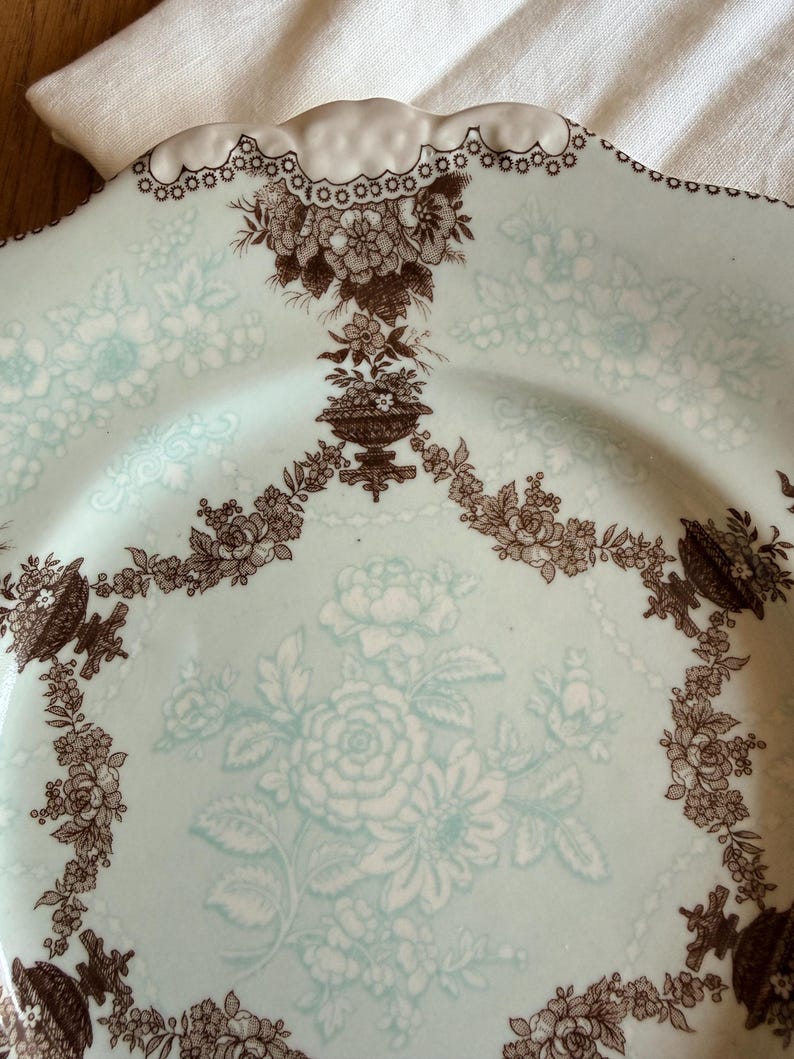 May include: A vintage, light blue plate with a floral design. The plate features a brown floral pattern around the edges and in the center. The rim has a scalloped edge with a white trim. The plate is likely made of ceramic.