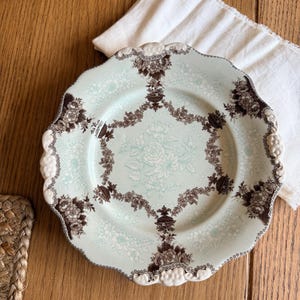 May include: A vintage, scalloped-edge plate with a light blue background and a floral pattern. The plate features a brown floral border and a central floral design. The plate is on a wooden surface with a white cloth.