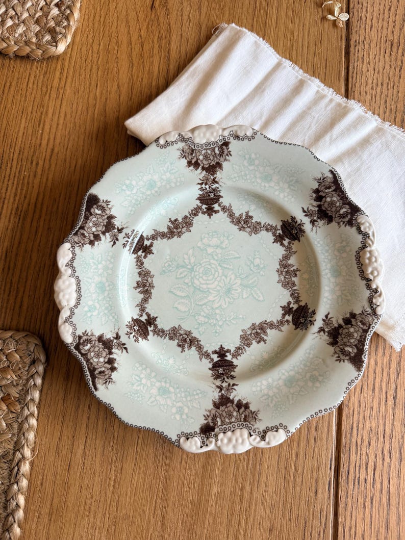 May include: A vintage, scalloped-edge plate with a light blue background and a floral pattern. The plate features a brown floral border and a central floral design. The plate is on a wooden surface with a white cloth.