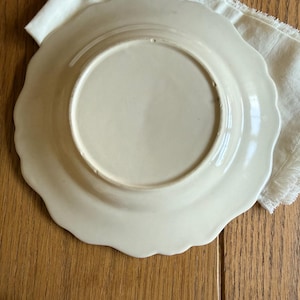 May include: Off-white ceramic plate with a scalloped edge, resting on a white linen napkin. The plate's underside is visible, showing a smooth, glossy surface. The background is a wooden surface.