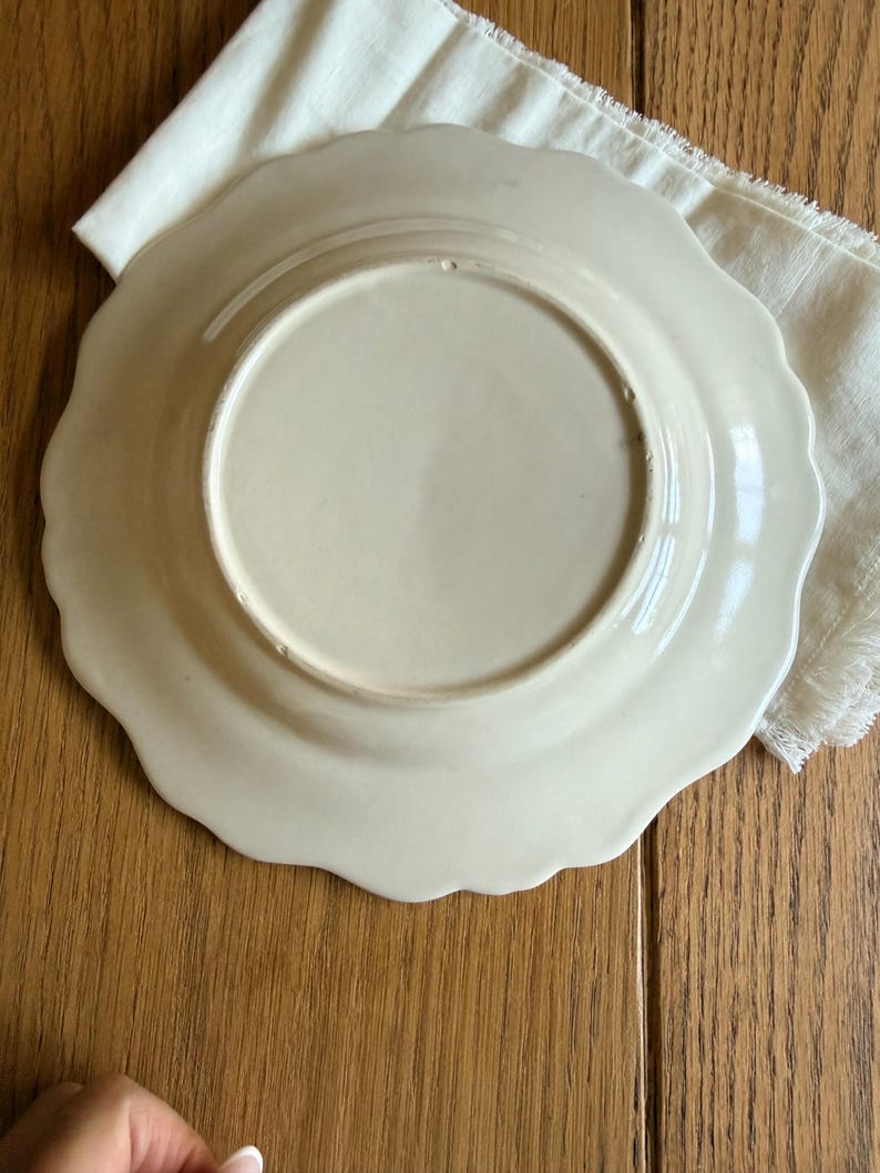 May include: Off-white ceramic plate with a scalloped edge, resting on a white linen napkin. The plate's underside is visible, showing a smooth, glossy surface. The background is a wooden surface.