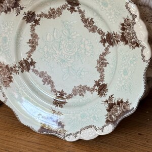 May include: A vintage porcelain plate with a scalloped edge. The plate features a light blue background with a floral pattern in white and brown. The rim is decorated with a brown and white floral design.
