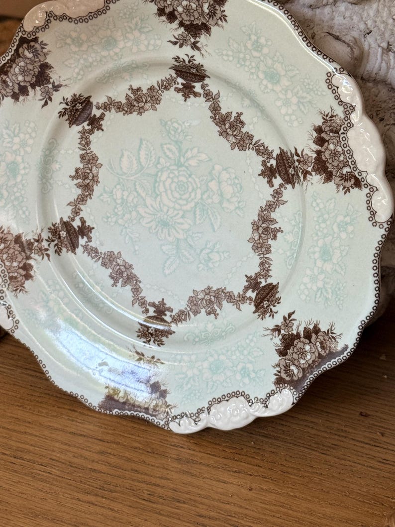 May include: A vintage porcelain plate with a scalloped edge. The plate features a light blue background with a floral pattern in white and brown. The rim is decorated with a brown and white floral design.