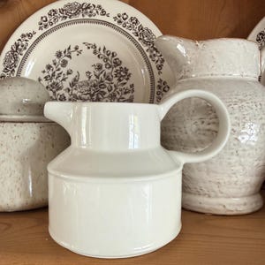 May include: A white ceramic pitcher with a handle and spout, positioned in front of a floral patterned plate and a textured white pitcher. The pitcher is a classic design, suitable for serving beverages. The background includes other ceramic pieces.
