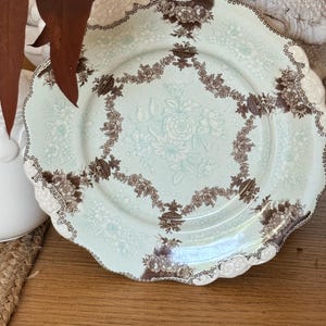 May include: A decorative plate with a scalloped edge. The plate features a light blue background with a floral pattern in white and brown. The rim is white with a brown dotted border. A brown floral garland encircles the center.