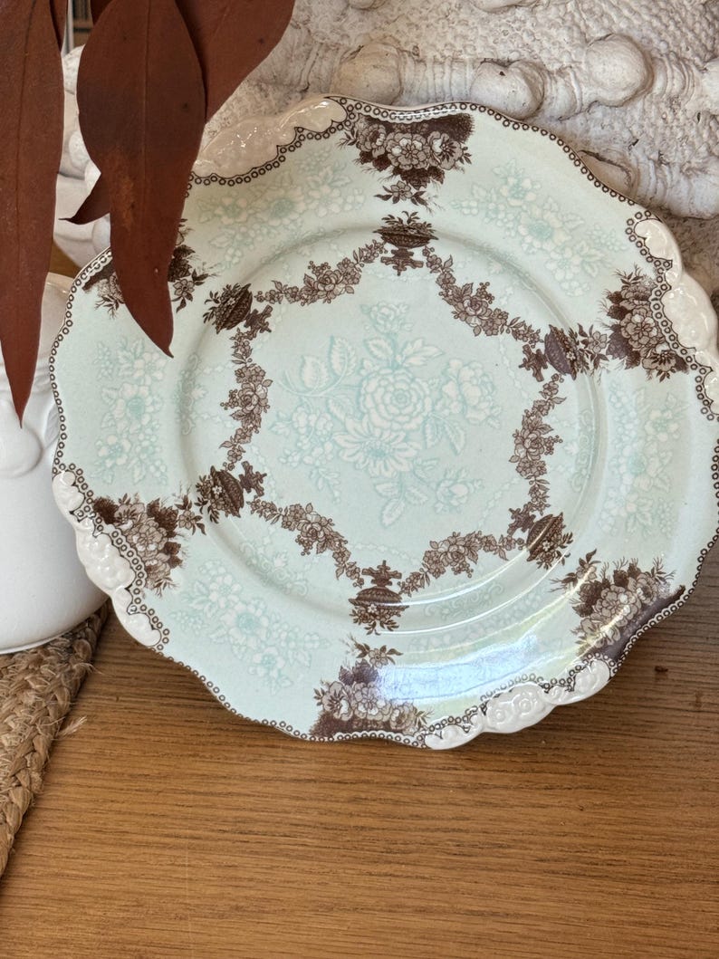 May include: A decorative plate with a scalloped edge. The plate features a light blue background with a floral pattern in white and brown. The rim is white with a brown dotted border. A brown floral garland encircles the center.