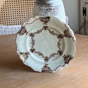 May include: A vintage porcelain plate with a scalloped edge. The plate features a light blue background with a brown floral pattern around the rim and center. The plate is displayed on a light wood surface.